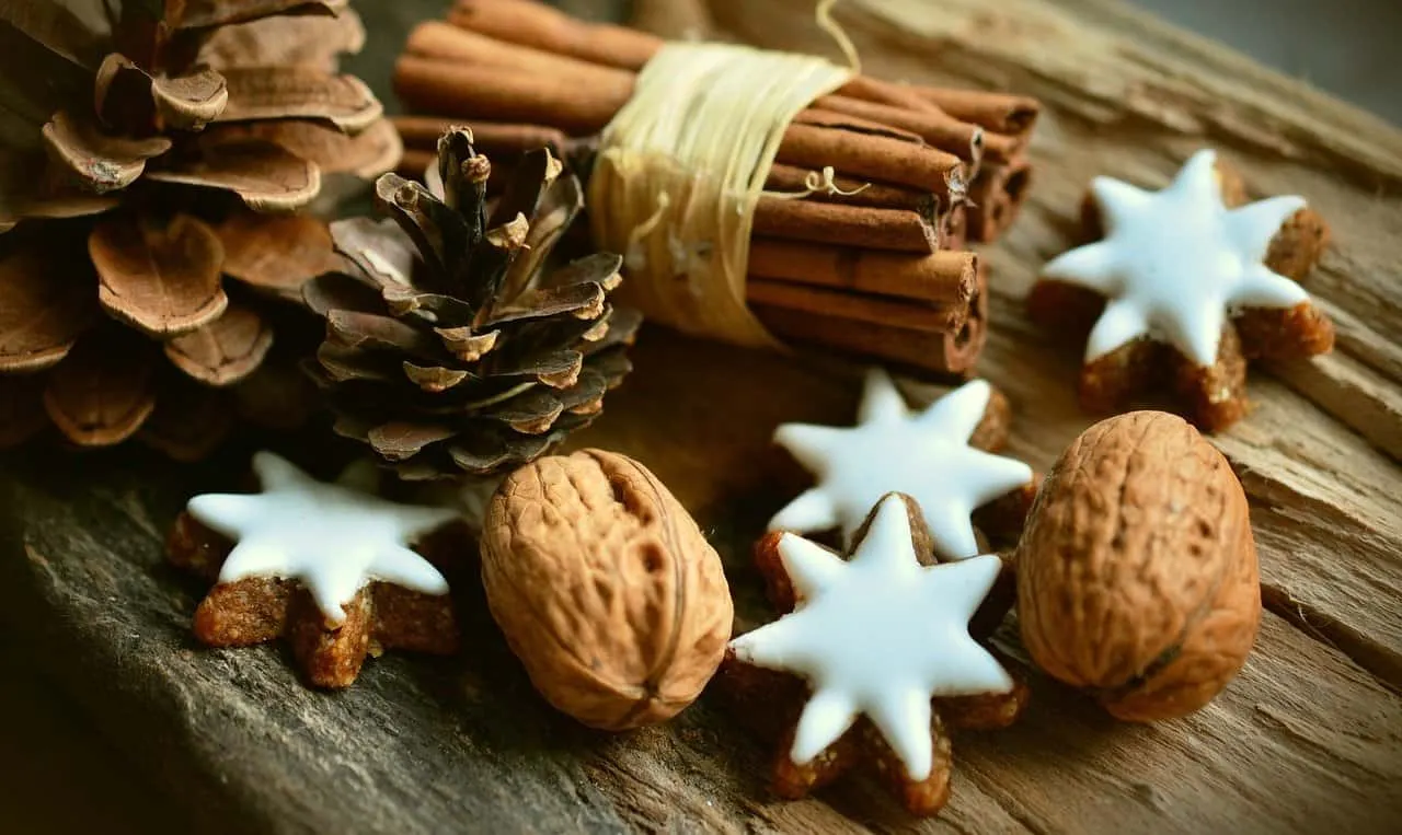 Sprigs of cinnamon, star-shaped cookies with white icing, walnuts, and pinecones arranged on rustic wooden surface for holida